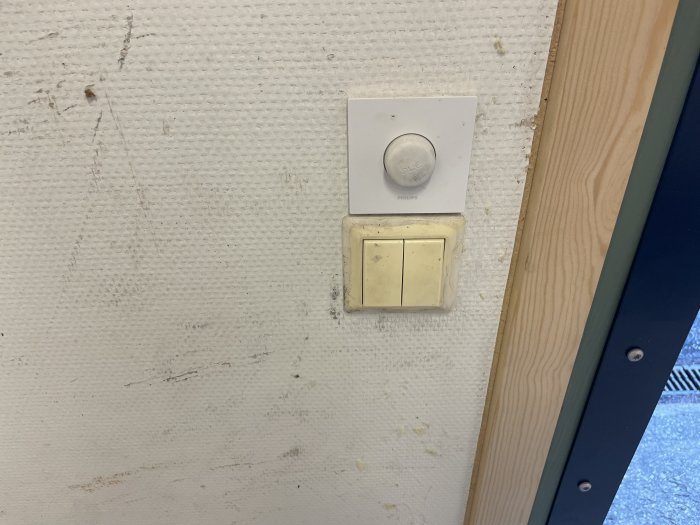 En väggmonterad Philips Hue smart-ljusströmbrytare ovanför en traditionell ljusbrytare på en smutsig vägg.