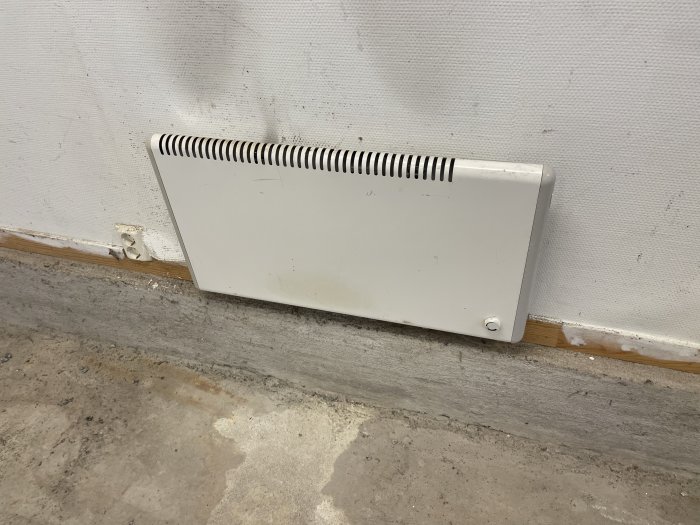 Vit vägghängd elradiator som står på ett dammigt betonggolv mot en vävtapetserad vägg.