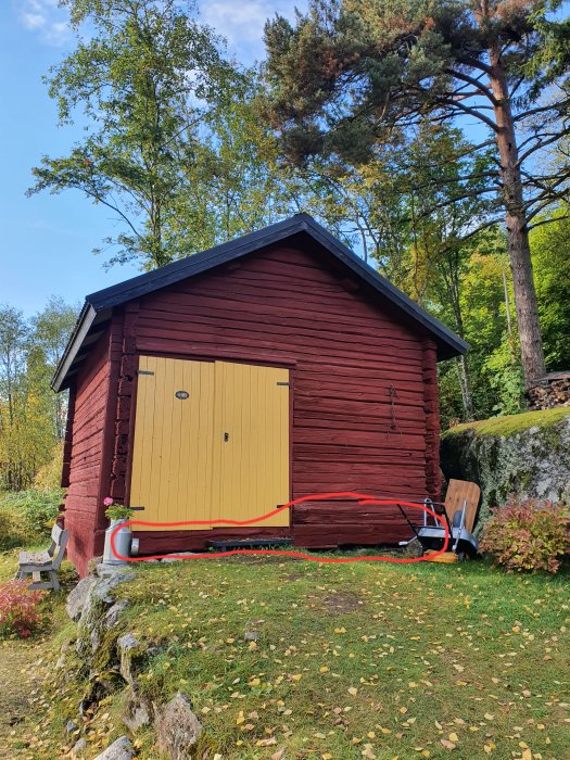 Röd timmerstuga med gul dörr, sladdvinda och trädgårdsverktyg utomhus.