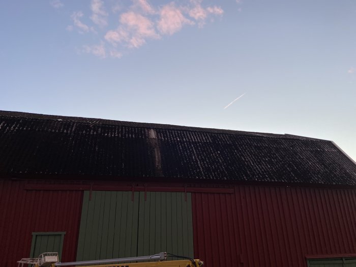 Ett rött och grönt lagerhus med mörkt tak i skymningen under en himmel med rosa moln och en flygplansstrimma.