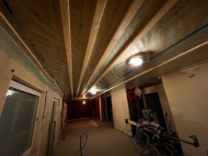 Byggarbetsplats med plasttak som är tejpade vid skarvarna, synliga takstolar och en taklampa.