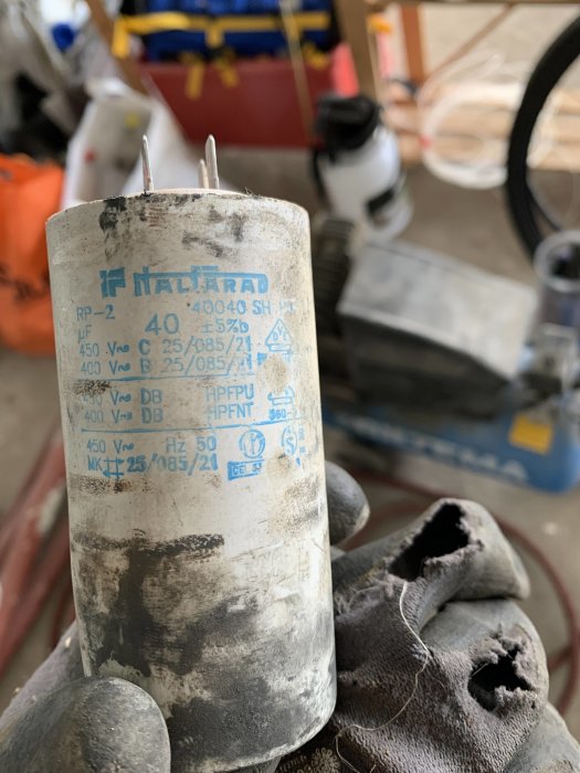 Närbild på en kondensator hållen av en person med slitna verktygshandskar i en verkstad.