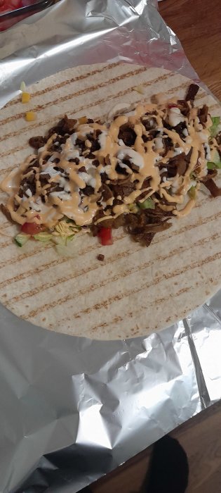 Öppen kebabrulle med grönsaker, kebabkött och vitlökssås på folieark.