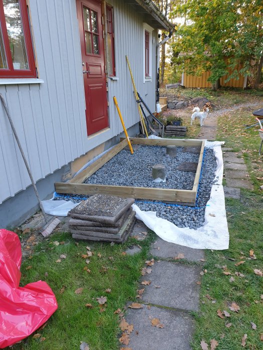 Pågående byggprojekt med halvfärdigt träramverk och grus på en trädgårdsgång bredvid ett hus, verktyg lutade mot väggen.