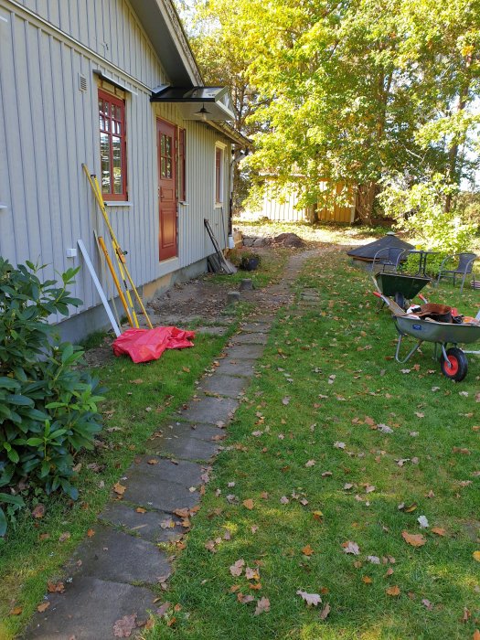 En stenlagd trädgårdsgång med arbetsmaterial och verktyg vid en husvägg, omgiven av grönska och höstlöv.