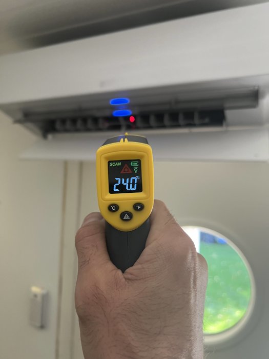 En hand håller en IR-termometer som visar 24,0 grader Celsius riktad mot en luftkonditioneringsenhet.