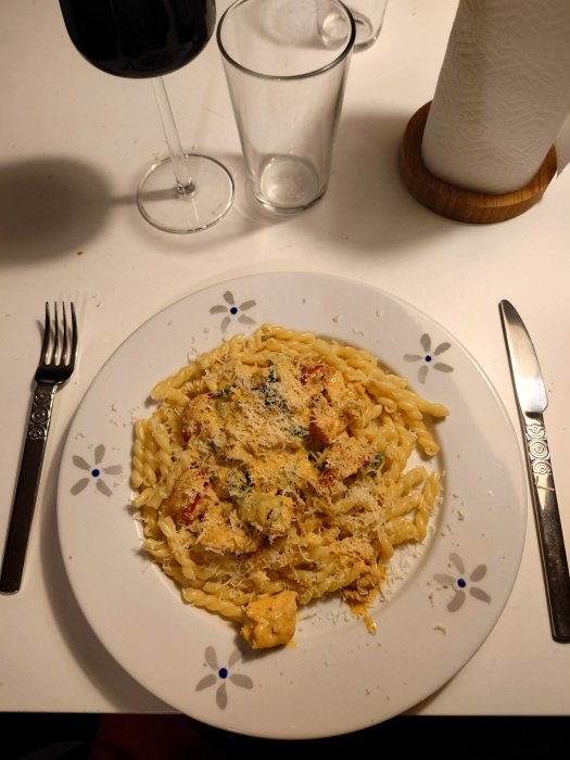 Tallrik med krämig curry kycklingpasta och riven ost, omgiven av bestick, glas och vinglas.