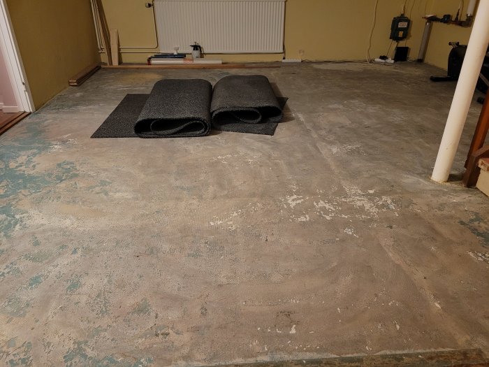 Opreparerat golv med två ihoprullade filtmattor i ett tomt rum under en renoveringsprocess.