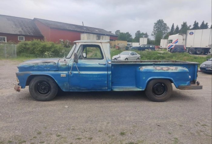 Blå pickup från 1977 med patina, parkerad utomhus med lantlig byggnad i bakgrunden.