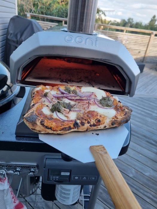 Nybakad pizza på en spade framför en Ooni Karu 12 pizzaugn på ett utomhuskök.