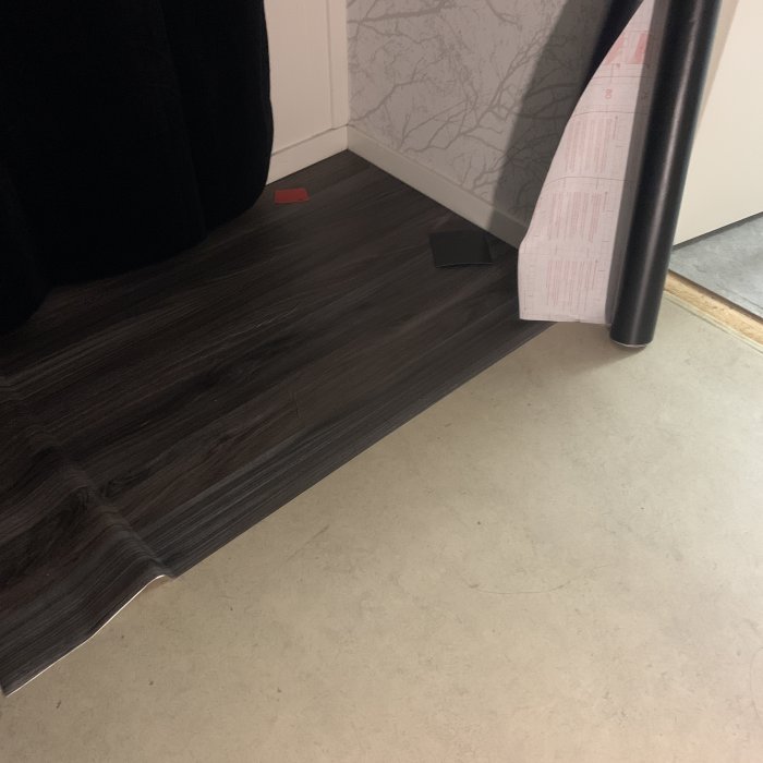 Mörk laminatgolv övergång till ljust betonggolv med svart dörrstopp och rullat papper i hörnet.