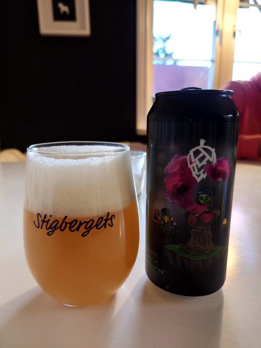 Ölglaset Stigbergets fyllt med öl, bredvid en burk Omnipollo med färgrik etikett på ett bord.