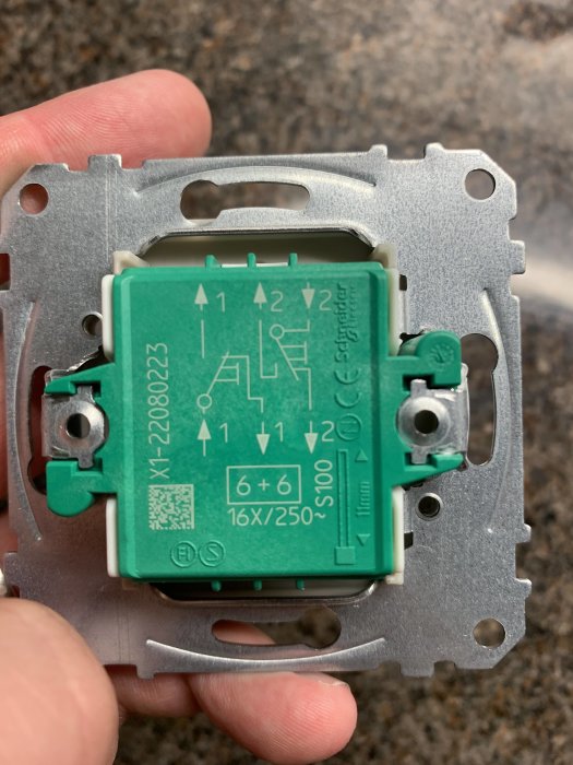 Hand håller en grön elektrisk strömbrytarmodul med schematisk märkning för installation.