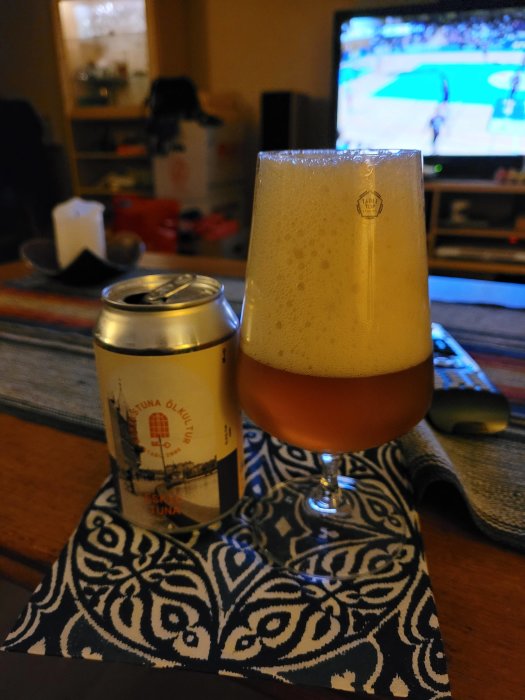 En burk Golden Ale från Eskilstuna Ölkultur bredvid ett glas fylld med öl, med en sportmatch i bakgrunden.