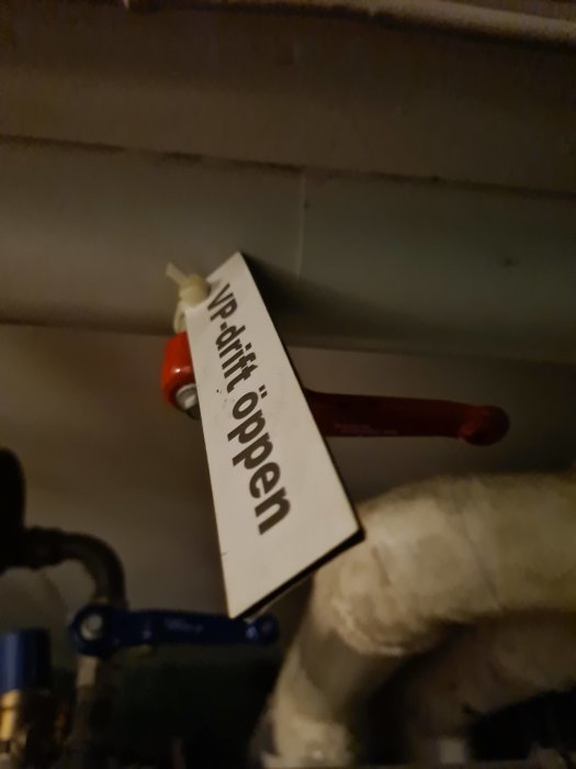 Etikett med texten "Vid drift öppen" som hänger på ett rör med ventiler i en teknisk installation.