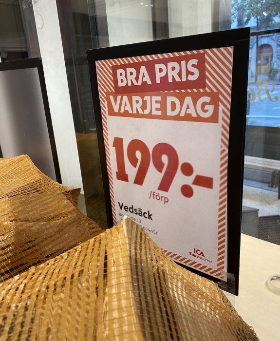 Skylt med texten "BRA PRIS VARJE DAG 199:- i förp. Vedsäck" framför säckar med björkved.