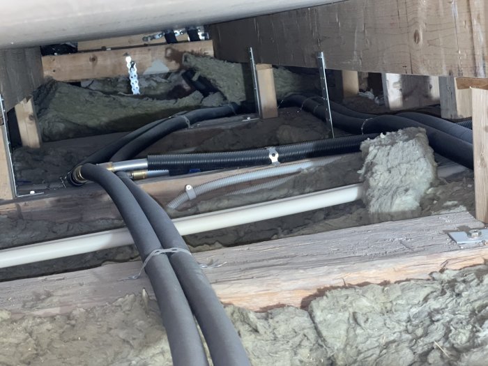 Vy under golv med olika rörinstallationer och isoleringsmaterial i ett pågående byggprojekt.