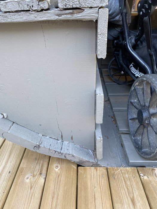 Skadad träpanel och fuktspärr på husgrund med sprickor och avskalad färg nära trätrall.