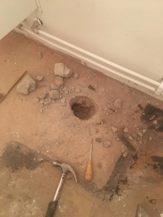 Golv med uppbrytning som avslöjar ett runt hål i betongen, omgivet av bråte och verktyg, inne i ett rum.