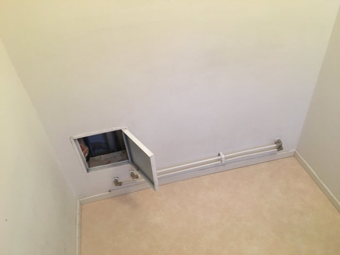 Vit vägg med en öppen inspektionslucka som visar rör, intill en radiator på en beige matta.