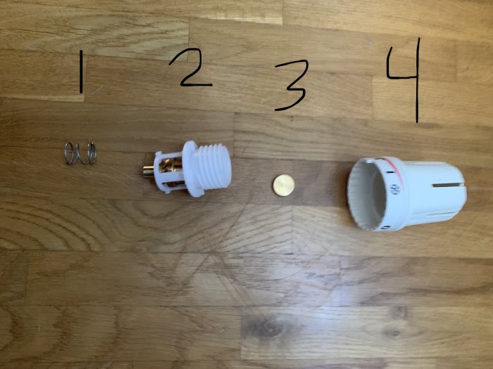 Två metallclips (1), vit lågenergilampa (2), mynt (3), och en vit plastadapter (4) på trägolv.