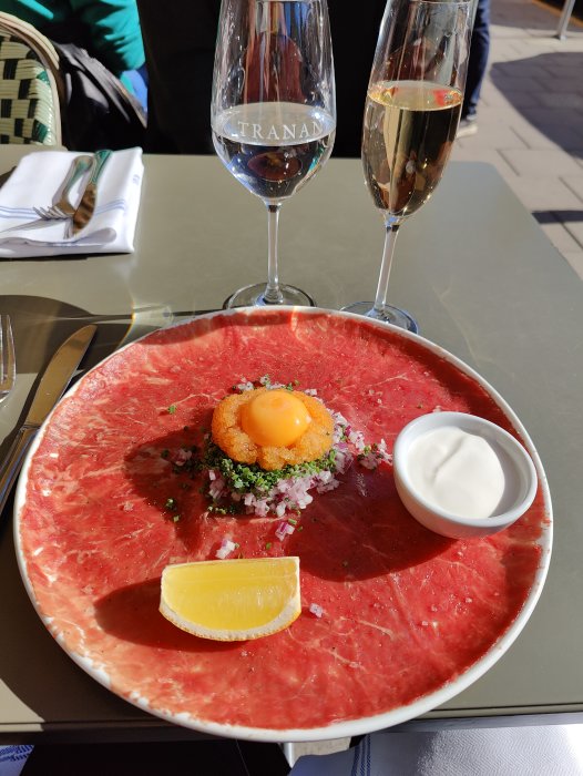 Carpaccio med äggula, citronklyfta och sås på en tallrik, vid en uteservering med två glas champagne.