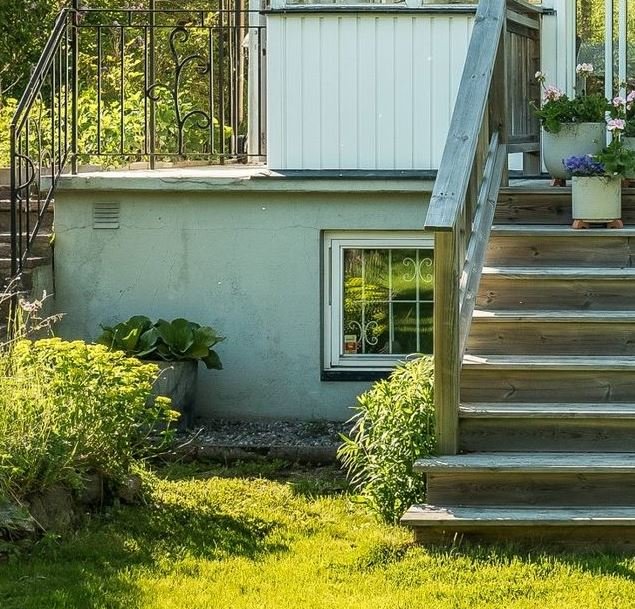 Trädgård utanför hus med källarfönster och trappa till altan, omgivet av grönska.