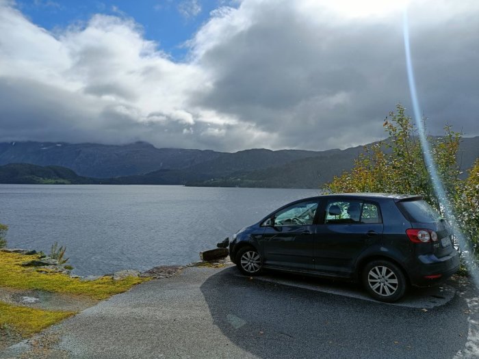 En mörkblå Golf Plus från 2009 parkerad vid en sjö med bergen och moln i bakgrunden.