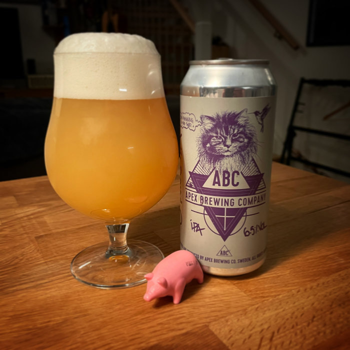 Ölglas med skummande öl bredvid en IPA ölburk från Apex Brewing och en liten rosa plastgris på ett träbord.