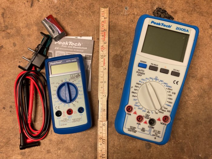 Två digitala multimetrar, PeakTech 1070 och 2005A, med testkablar och manual på en arbetsbänk.