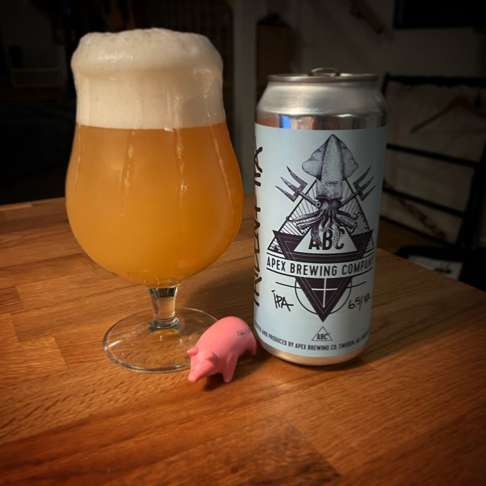 Fyllt ölglas med skum bredvid en burk från Apex Brewing Company och en rosa plastgris på ett bord.