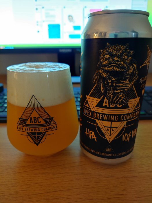Ölglas fyllt med ljus öl framför en dator, och en burk Apex Brewing Company öl.