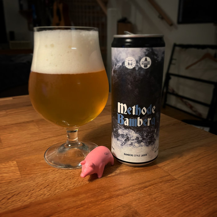 Ölglas med skum och burk märkt "Methode Bamberg", samt en liten rosa leksaksgris på ett bord.