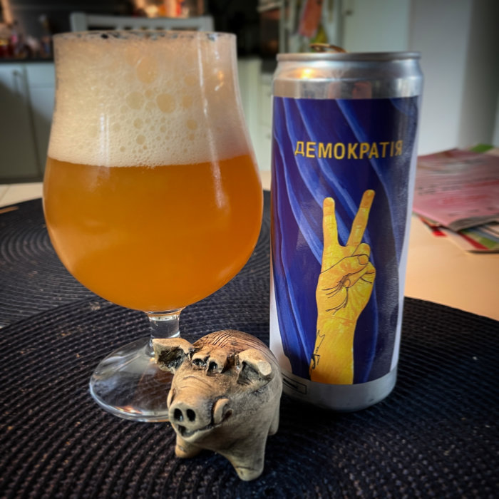 Ett glas med öl bredvid en ölburk med "fredstecken"-motiv och en liten trägrisfigur i förgrunden.