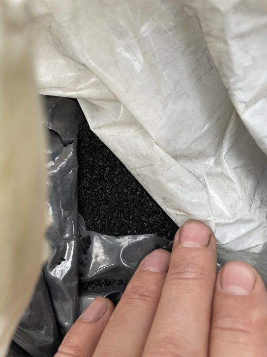 En hand öppnar en vit säck för att visa svarta byggmaterialgranulat inuti.