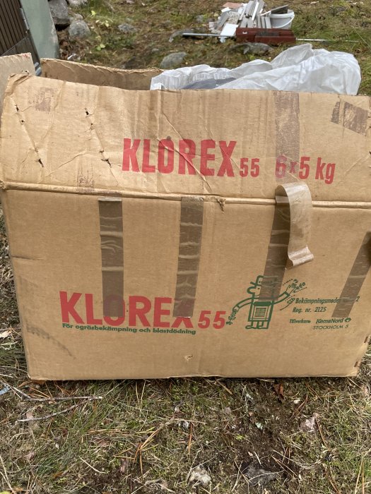 Nöten brun kartong med texten "KLOREX 55 6x5 kg" för ogräsbekämpning och bladdelning, utomhus.