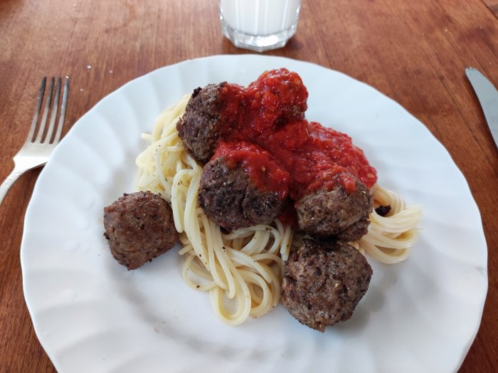 Spaghetti med köttbullar toppade med tomatsås på en vit tallrik, gaffel och glas med mjölk i bakgrunden.