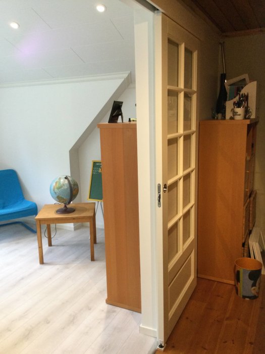 Vitmålad dörr halvöppen in till ett rum med snedtak, träbyrå, blå stol och ett skrivbord.