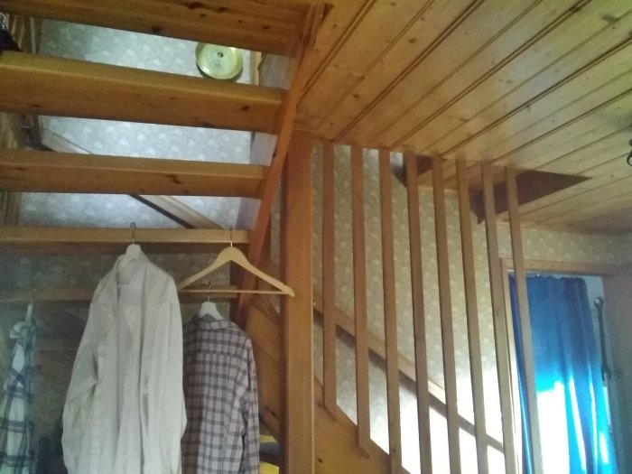 Interiörbild av ett träloft med räcke och kläder på galgar.