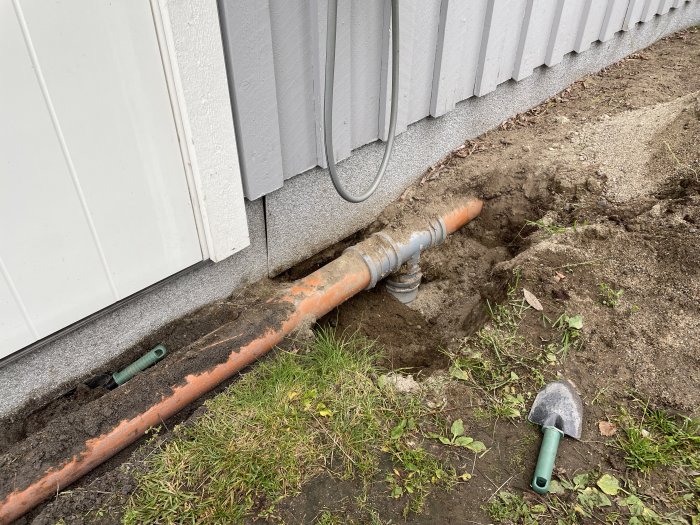 Grävd grop vid husgrund som visar en exponerad orange avloppsrör delvis täckt av jord och en handspade i förgrunden.