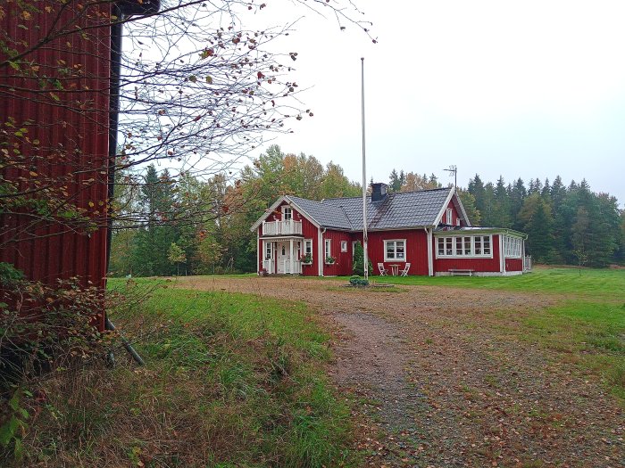 Rött trähus med vita knutar och en veranda, omgivet av grönt gräs, träd och en grusgång, under en molnig himmel.