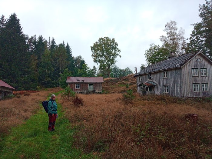 Vandrare framför gammalt hus och lada i landsbygd med torrt gräs och skog i bakgrunden.
