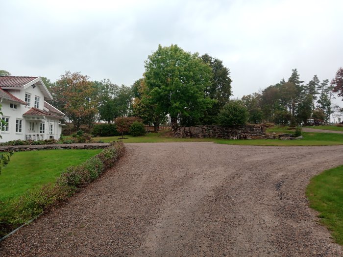 Vit villa vid en grusväg med välskött gräsmatta och stenmur, omgiven av träd.