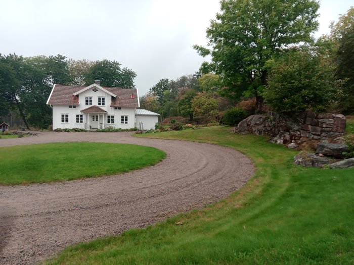 Vit villa med röda takpannor vid en gårdsplan med slingrande grusväg, gräs och stenmur i skymundan.