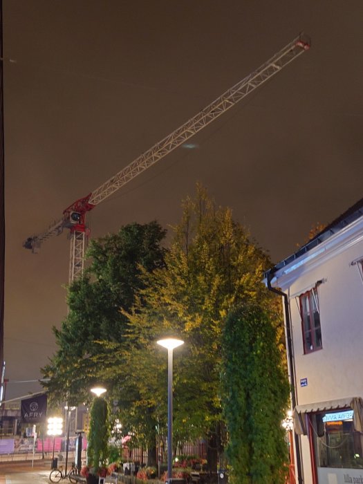 Nattbild av en byggnadskran ovanför träd och byggnader i en stadsmiljö.