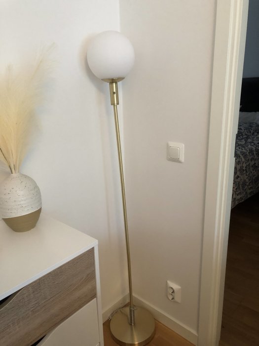 Guldfärgad golvlampa med rund vit skärm i hörn bredvid vit byrå och ljus vägg.