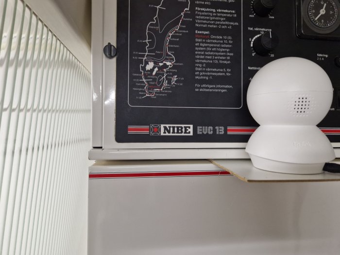 NIBE värmesystem med kontrollpanel och en IP-kamera på toppen ovanför en vit radiator.