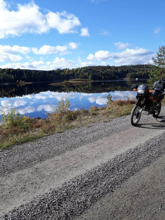 Motorcykel parkerad vid en stilla sjö omgiven av skog och spegelblankt vatten på en solig höstdag.