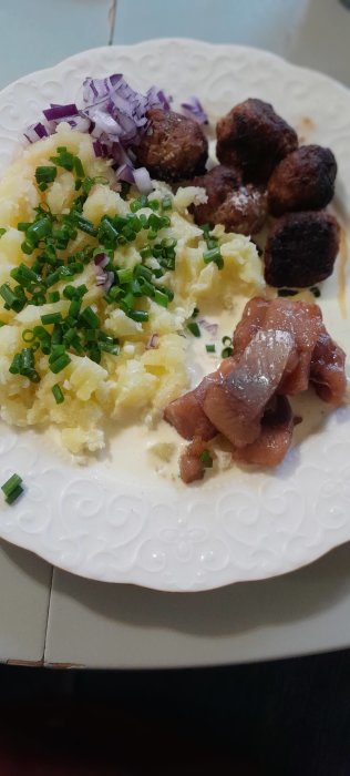 Tallrik med potatismos, köttbullar, inlagd sill och hackad rödlök, toppat med gräslök.