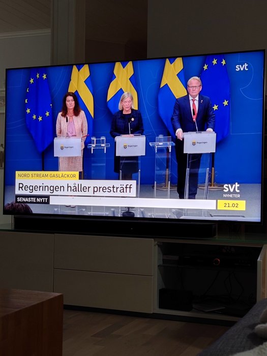 TV-skärm visar nyhetsprogram med tre talare framför svenska flaggor och EU-flaggor.